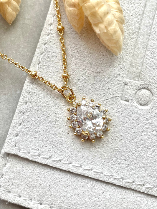 A gold-plated cubic zirconia teardrop pendant necklace with a chain, displayed on a white textured surface next to a white card and a shell.