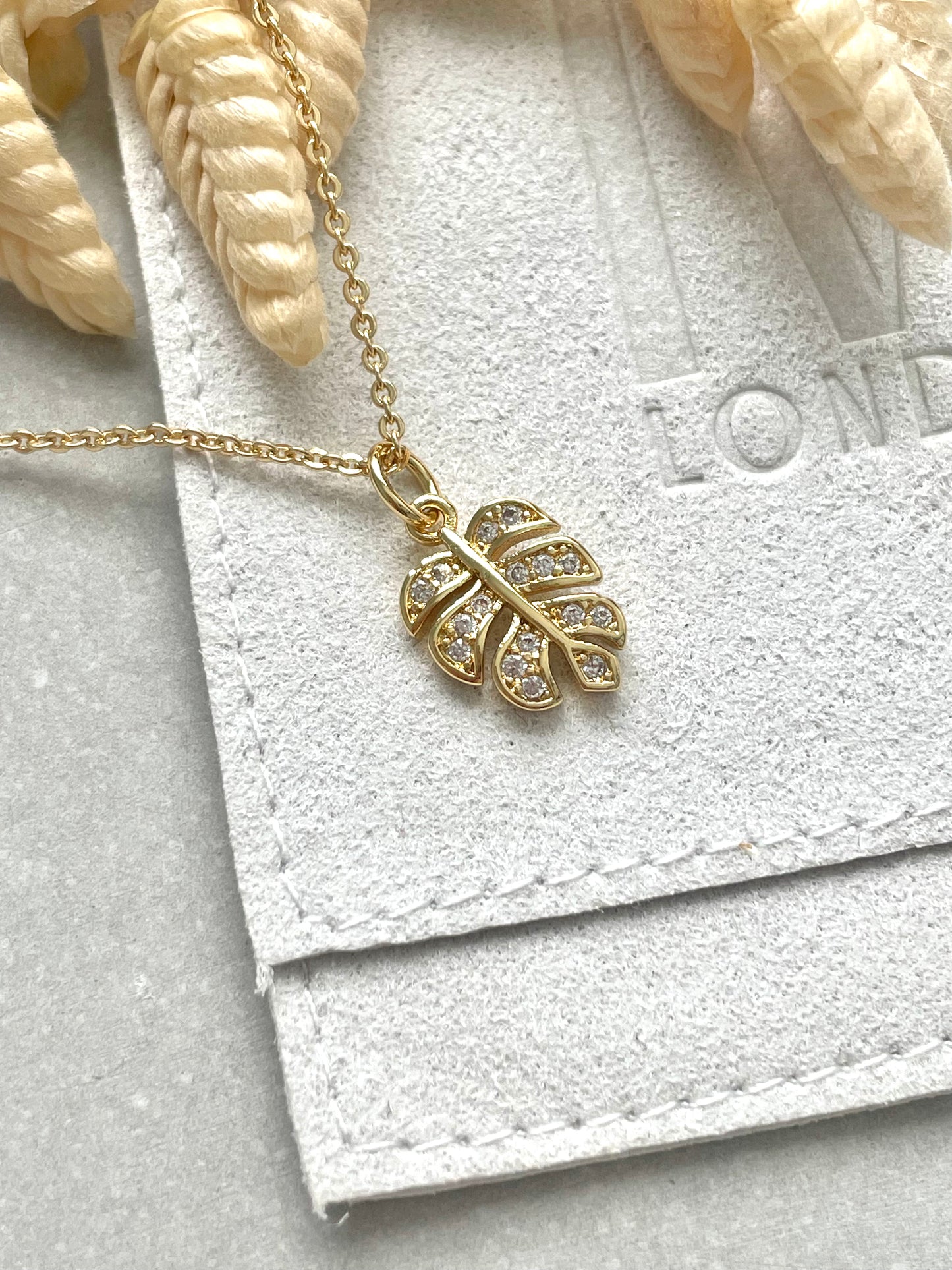 An 18k gold plated cubic zirconia monstera leaf necklace with a chain and a trigger clasp, displayed on a textured background.