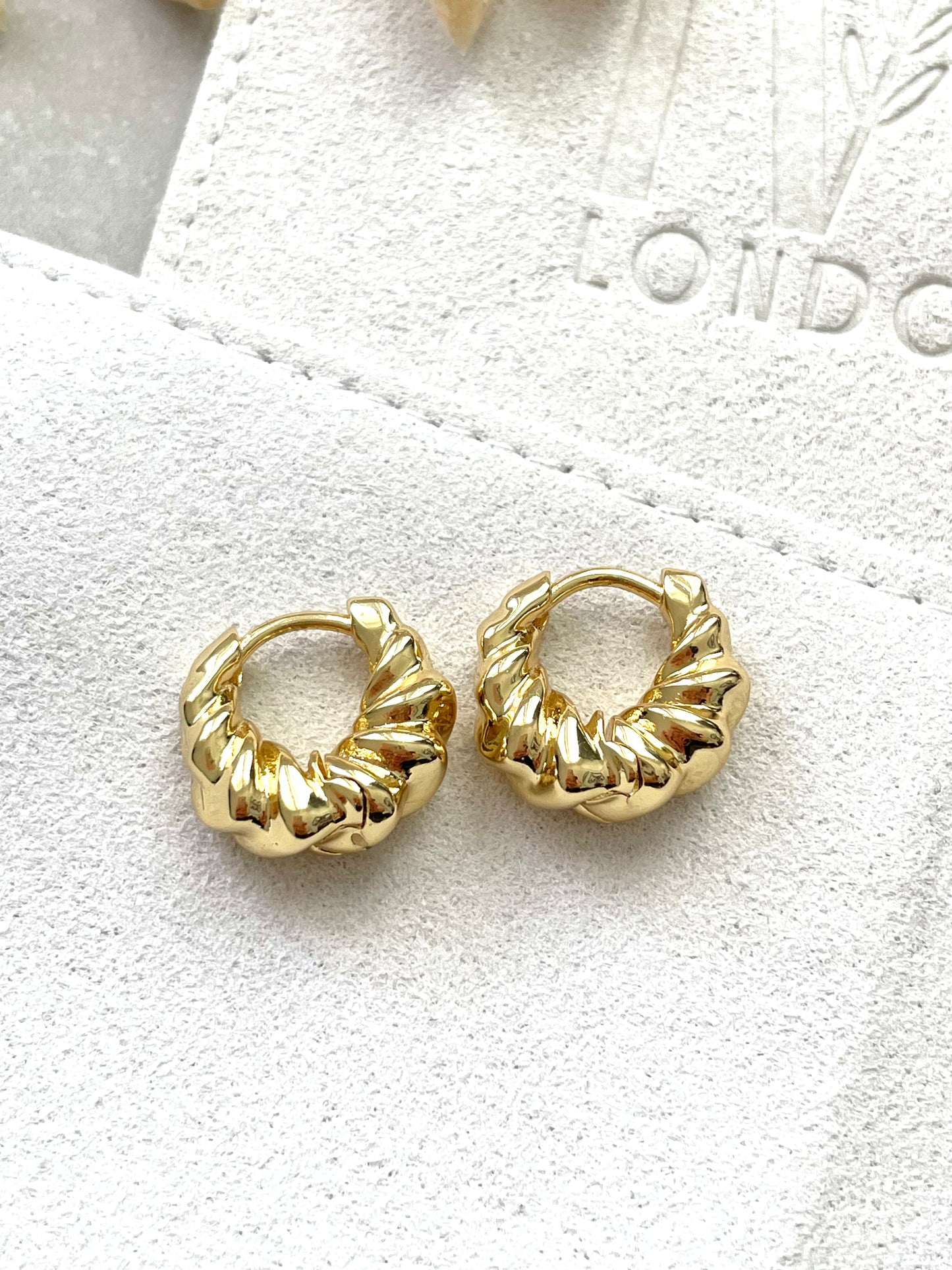 A pair of gold-plated hoop earrings with a ripple effect design, displayed on a white fabric surface.