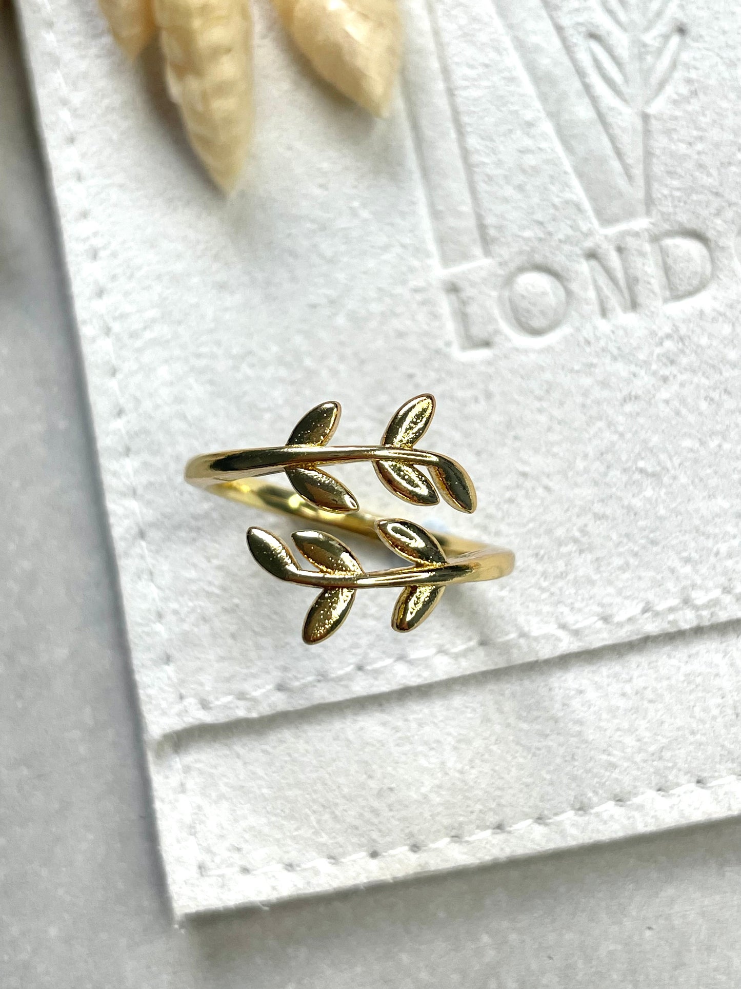 A gold-plated olive leaf ring with two leaf-shaped settings, placed on a white fabric surface.