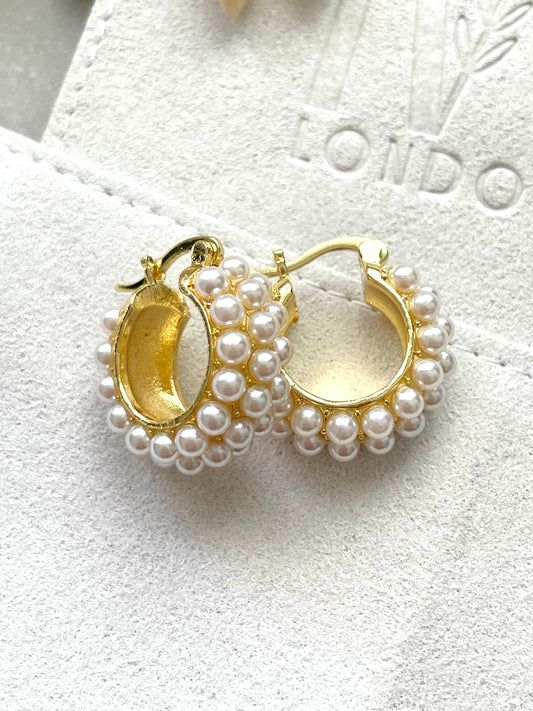 A pair of gold-plated pearl earrings with a textured surface and a brand logo in the background.