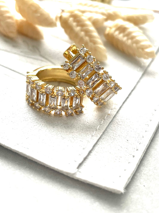 A pair of gold-plated baguette-shaped cubic zirconia earrings displayed on a white surface with a woven textile in the background.