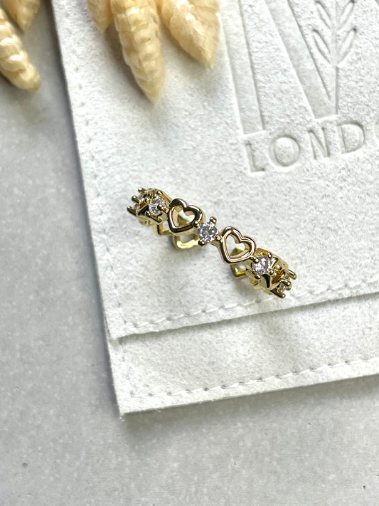 A gold-plated cubic zirconia ring with a heart shape design, placed on a white paper with a decorative object in the background.