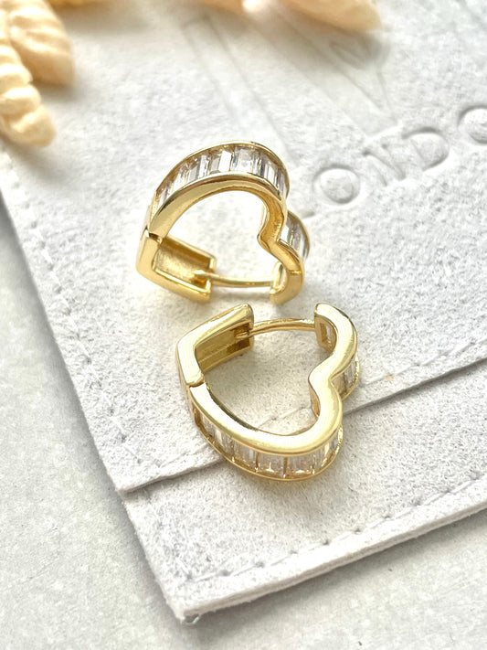 A pair of gold-plated, heart-shaped hoop earrings on a white textured surface.