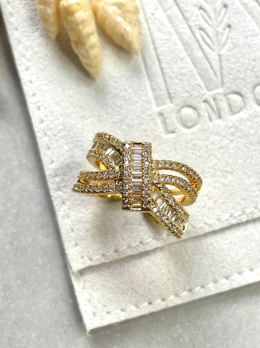 A gold-plated cubic zirconia layer ring with a layered design, resting on a soft surface, with a small piece of white textured material to the left.