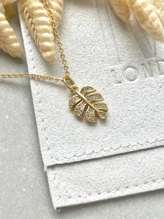 An 18k gold plated cubic zirconia monstera leaf necklace with a chain and a trigger clasp, displayed on a textured background.