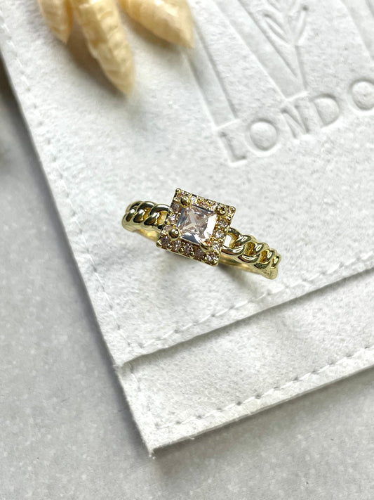 A gold plated cubic zirconia square cut stone ring displayed on a soft surface with a branded card in the background.