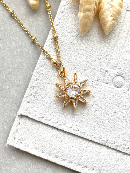 A gold plated cubic zirconia star flower necklace with a chain and a trigger clasp, displayed on a white textured surface.