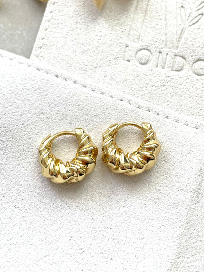A pair of gold-plated hoop earrings with a ripple effect design, displayed on a white fabric surface.