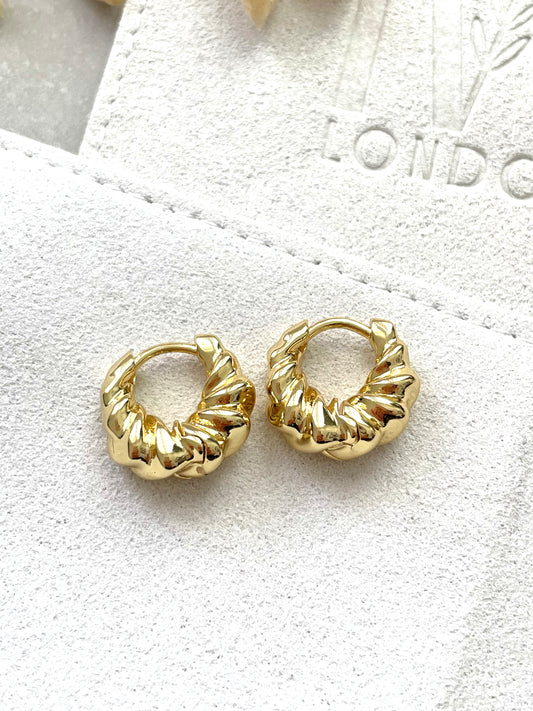 A pair of gold-plated hoop earrings with a ripple effect design, displayed on a white fabric surface.
