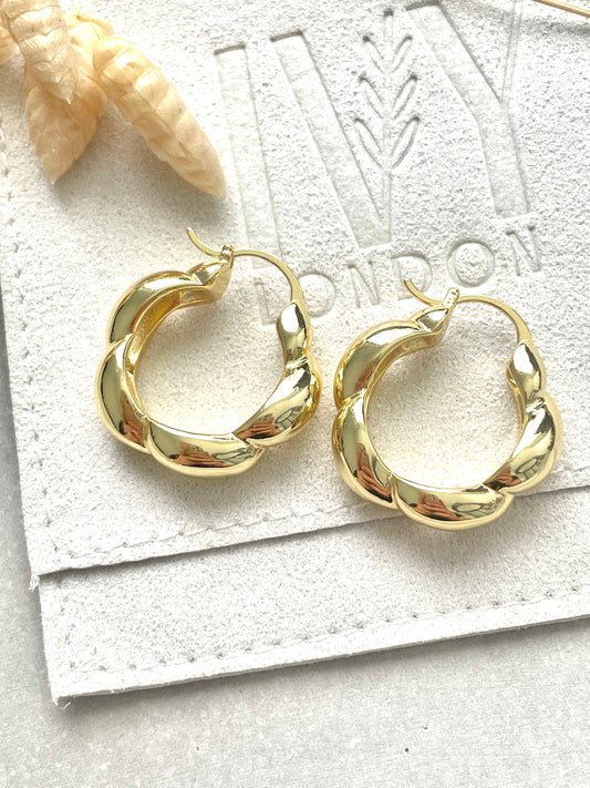 Gold plated twist earrings laid on a white surface with a cardboard in the background.