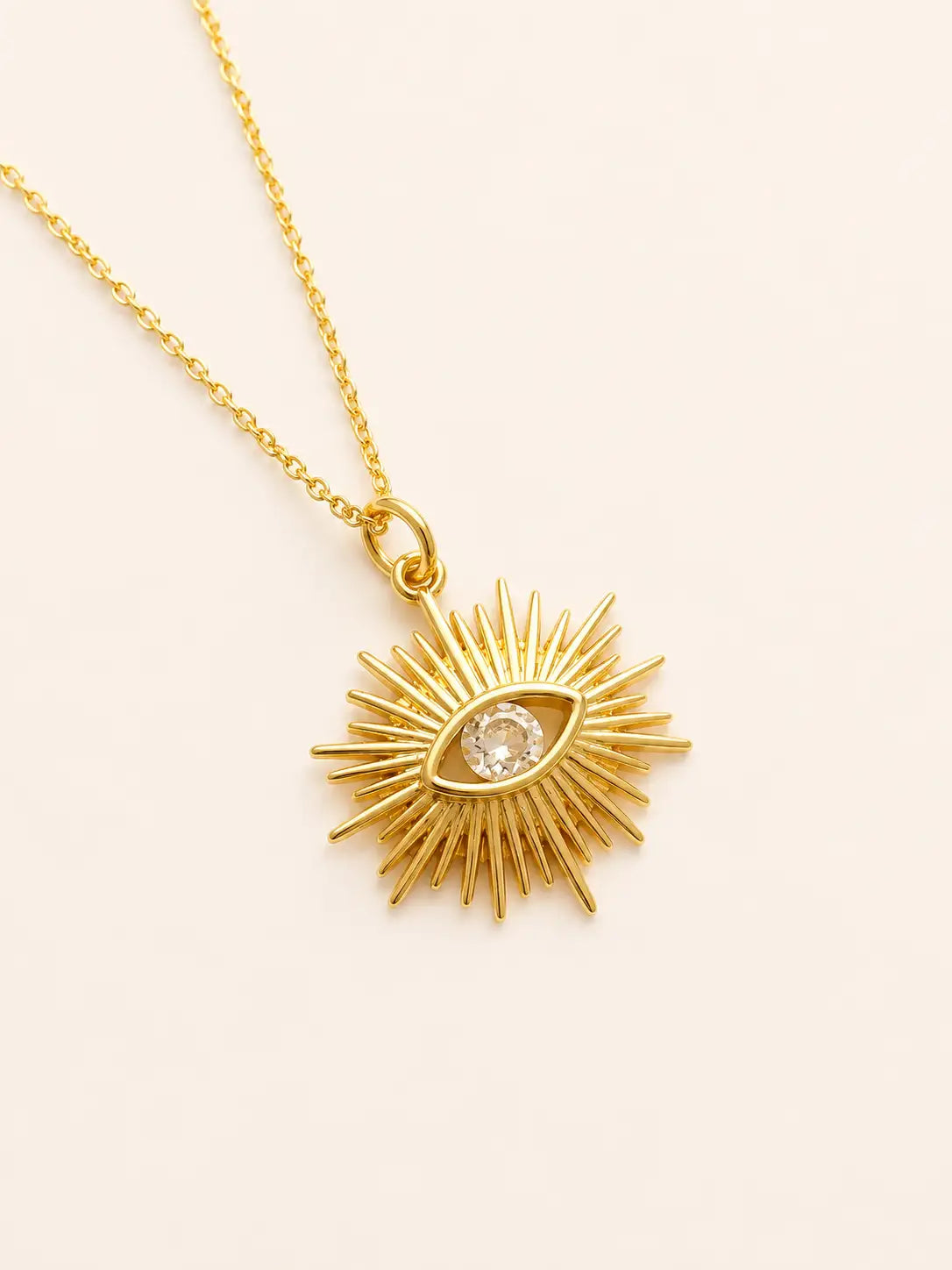 Gold necklace with a sunburst pendant featuring an eye design on a beige background