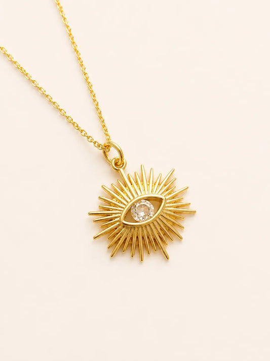 Gold necklace with a sunburst pendant featuring an eye design on a beige background